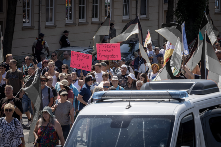 einige Menschen, die neben einem Polizeiauto herlaufen und Deutschlandfahnen schwenken, sowie Plakate mit der Aufschrift "unser Geld f&uuml;r unser Volk" und "Frieden, Freiheit, Remigration" halten