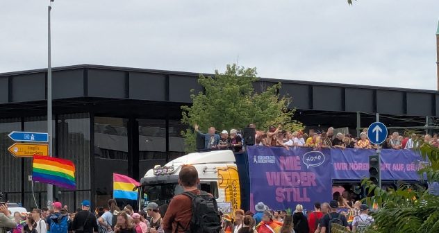 Demonstrierende des CSD Berlin, z.B. mit Regenbogenflagge, und ein Lkw, auf dem ebenfalls Demonstrierende stehen und an dem eine Plane mit dem CSD-Motto "Nie wieder still!" hängt.