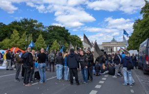 Junge Neonazis mit Deutschland-Fahnen bei einem Aufmarsch auf der Straße des 17. Juni. Hinter ihnen in der Ferne das Brandenburger Tor, neben ihnen weitere Teilnehmende, teilweise mit Friedens- und Russlandflaggen.