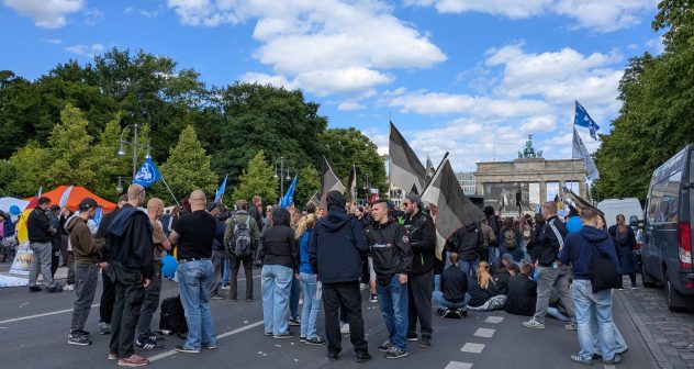 Junge Neonazis mit Deutschland-Fahnen bei einem Aufmarsch auf der Straße des 17. Juni. Hinter ihnen in der Ferne das Brandenburger Tor, neben ihnen weitere Teilnehmende, teilweise mit Friedens- und Russlandflaggen.